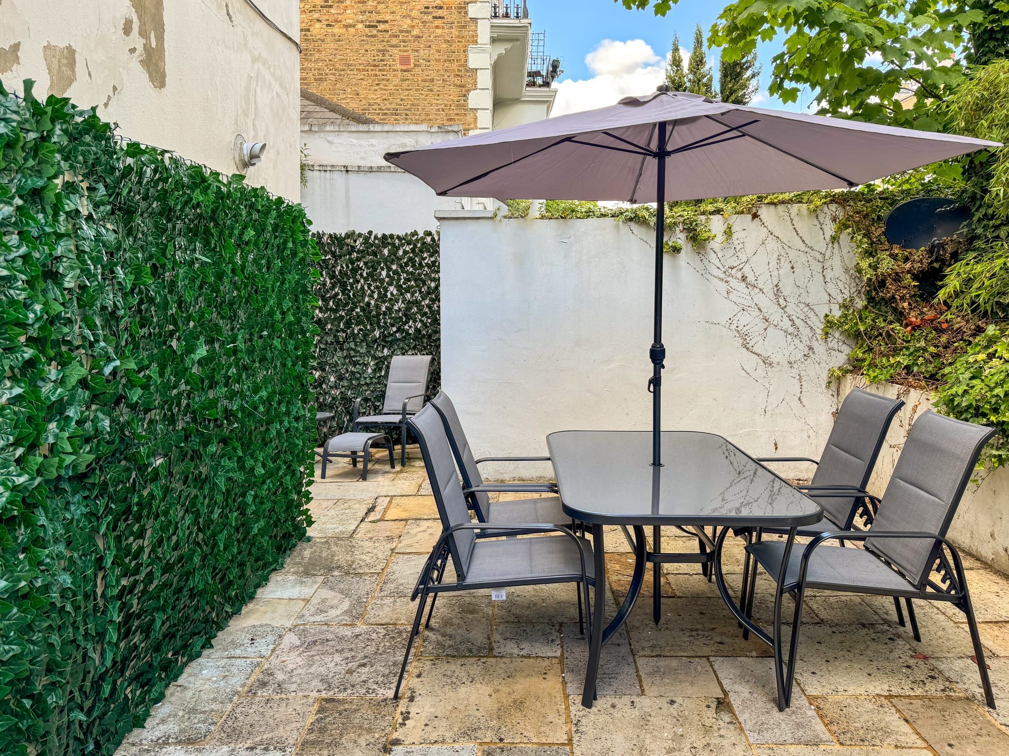 Eine Terrasse im Freien mit Tisch und Stühlen, einem großen Sonnenschirm und einer Hecke.