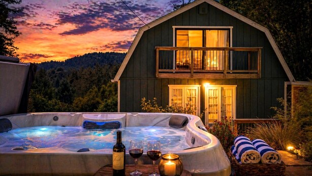 A two-story wooden cabin with a hot tub and a balcony, surrounded by trees and a sunset view. - MagicMountain~River Hideaway~View~Hot Tub~Dog~Summer Magic Retreat! EV Charge (Cazadero)