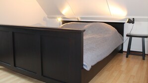 A bedroom with a dark wooden bed, a bedside table with a lamp, and a sloped ceiling with two wall-mounted lights. - Nice Holiday Home 200m. From the Beach (Wemeldinge)