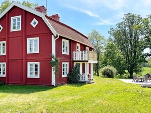 A red two-story house with a balcony, surrounded by a grassy lawn and trees.