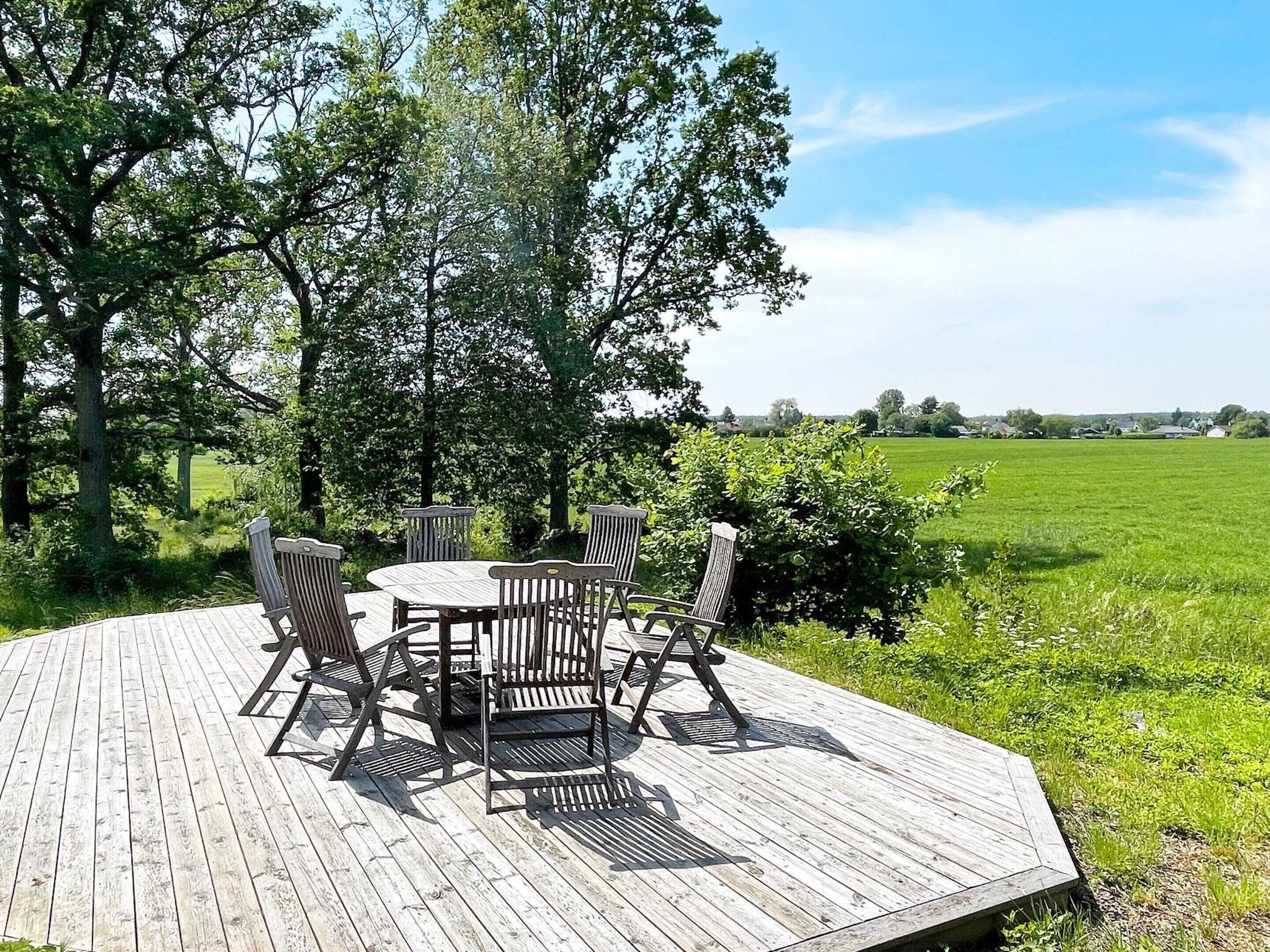 A wooden deck with a table and chairs, surrounded by greenery and a view of a field.