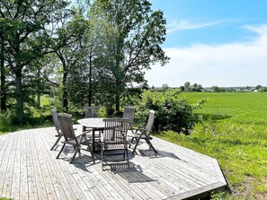 A wooden deck with a table and chairs, surrounded by greenery and a view of a field.