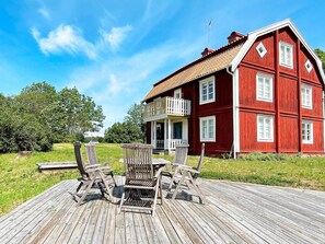 Ein rotes Holzhaus mit einem Balkon und einer Holzterrasse mit Stühlen, vor einem strahlend blauen Himmel.