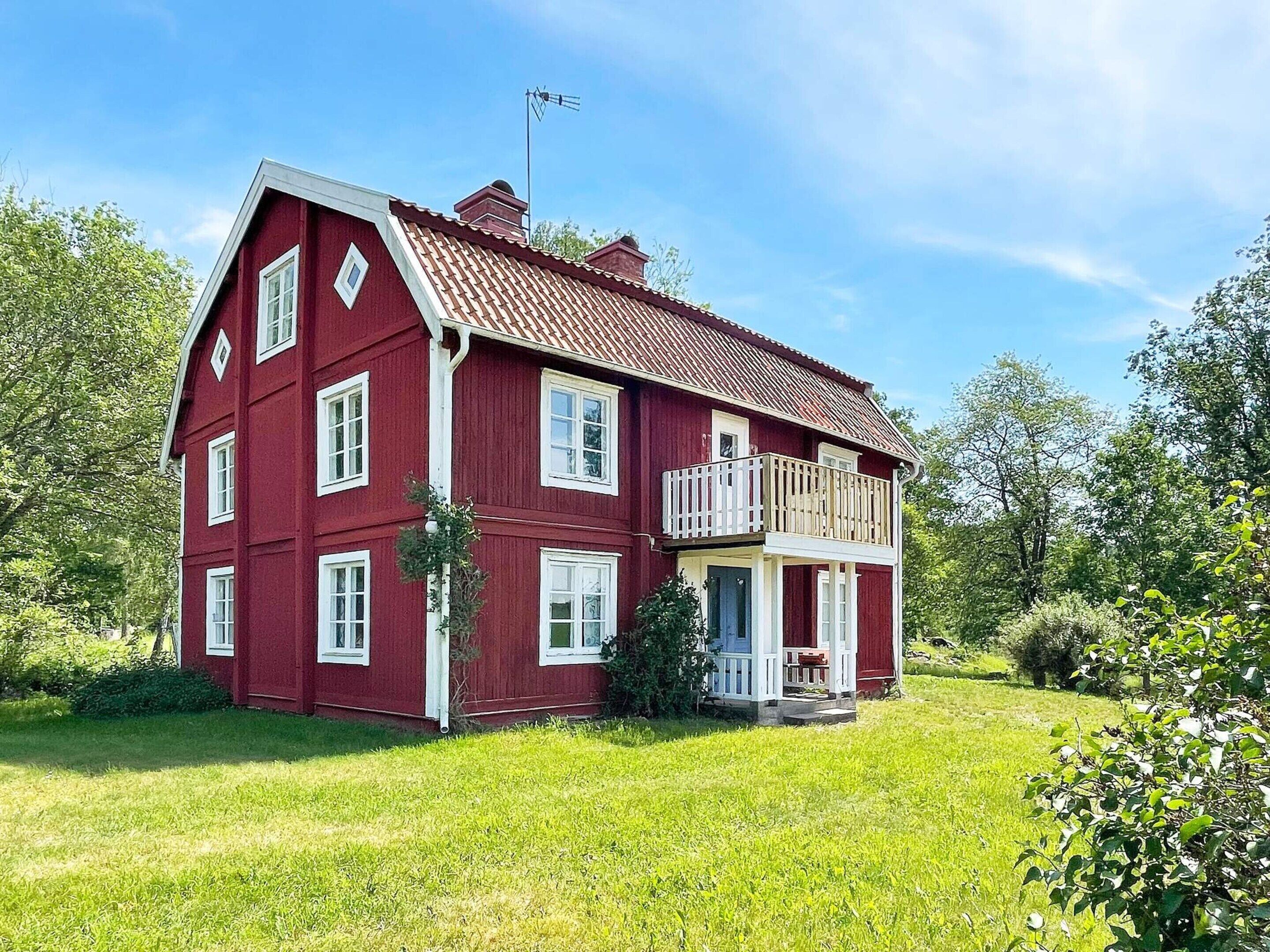 A two-story red house with a balcony, surrounded by greenery.