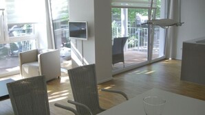 Ein modernes Zimmer mit einem großen Schiebeglasfenster, das zu einem Außenbereich führt, mit Korbmöbeln, einem weißen Tisch und einem flachbildfernseher an der Wand.