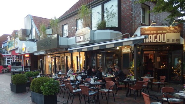 Ein Straßencafé mit Tischen und Stühlen, umgeben von Ziegelgebäuden mit verschiedenen Schildern.