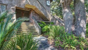A house with a wooden staircase leading to a front door, surrounded by lush greenery and a large tree. - Kiawah Exclusives | 4270 Mariners Watch (Johns Island)