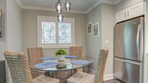 A dining area with a round glass table, woven chairs, a stainless steel refrigerator, and a window with blinds. - Kiawah Exclusives | 4270 Mariners Watch (Johns Island)