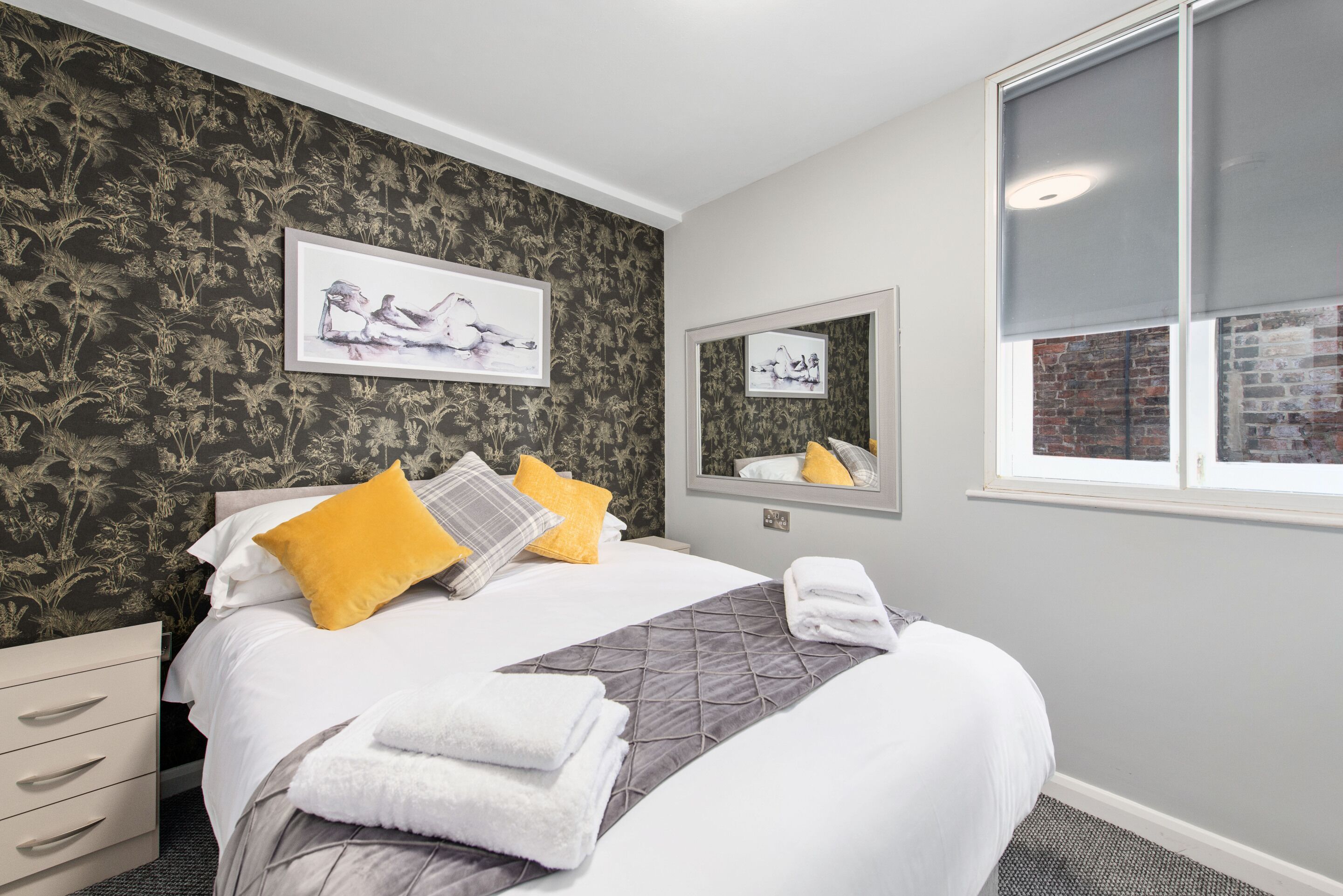 A neatly made bed with white bedding and a gray quilt, a yellow pillow, and a gray throw blanket. There are two framed pictures on the wall, a mirror above the bed, and a window with a view of a brick wall.