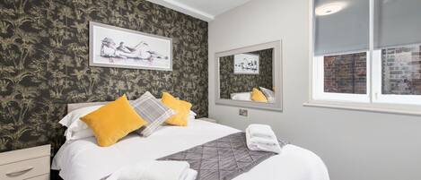 A neatly made bed with white bedding and a gray quilt, a yellow pillow, and a gray throw blanket. There are two framed pictures on the wall, a mirror above the bed, and a window with a view of a brick wall.