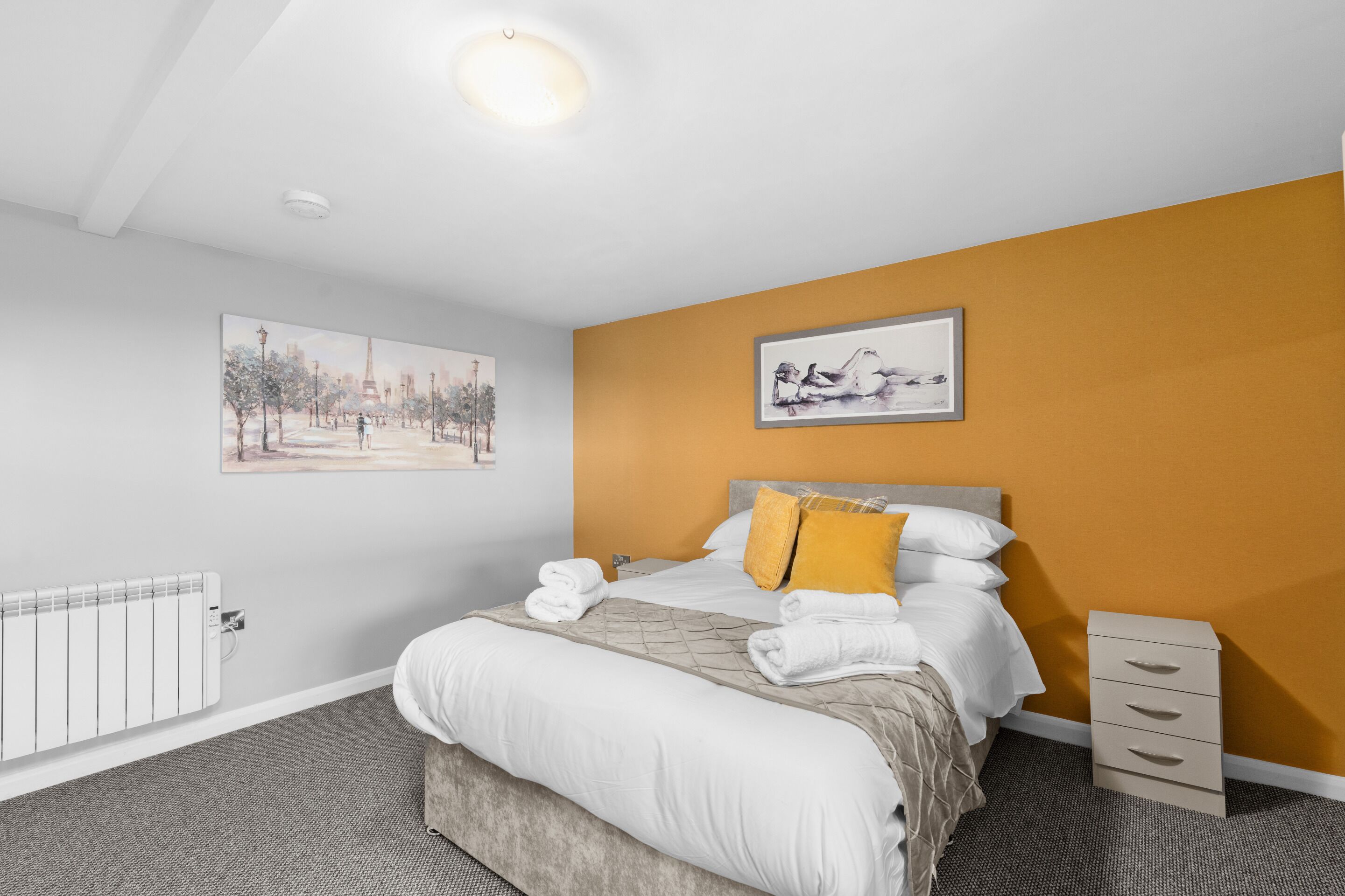 Une chambre d’hôtel avec un lit, un radiateur, une table de chevet et deux tableaux encadrés au mur.