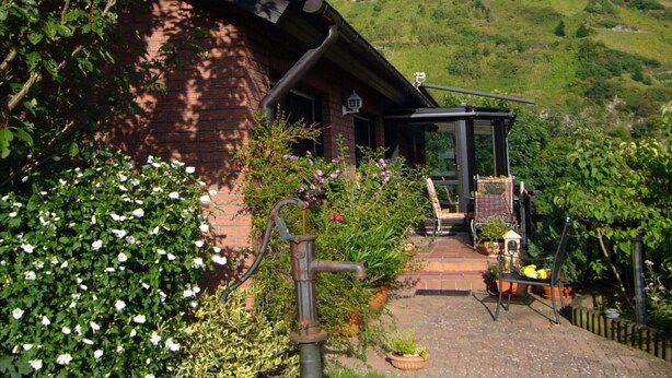 En rustik bygning med en murstensfacade, en overdækket veranda med et bord og stole, og en have med forskellige planter og blomster.