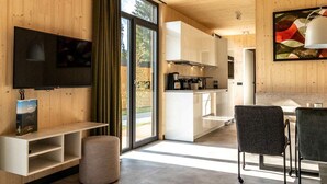 A modern interior with a flat-screen TV, a dining area with chairs, a kitchen with white cabinets, and a sliding glass door leading to a garden. - Modern Holiday Home With sun Shower (Winterberg)