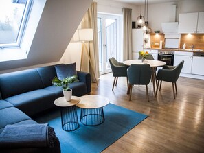 A modern living room with a sofa, a dining table with chairs, a kitchen area, and a large window with curtains.