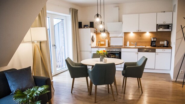 A modern living room with a sofa, a coffee table, and a dining area with chairs and a table. There is a kitchen in the background with white cabinets and a microwave. - Narrow Luzin Modern Retreat (Feldberger Seenlandschaft)