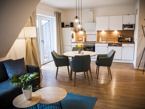 Una sala de estar moderna con sofá, una mesa de centro y un comedor con sillas y una mesa. Se observa una cocina al fondo con armarios blancos y un microondas.