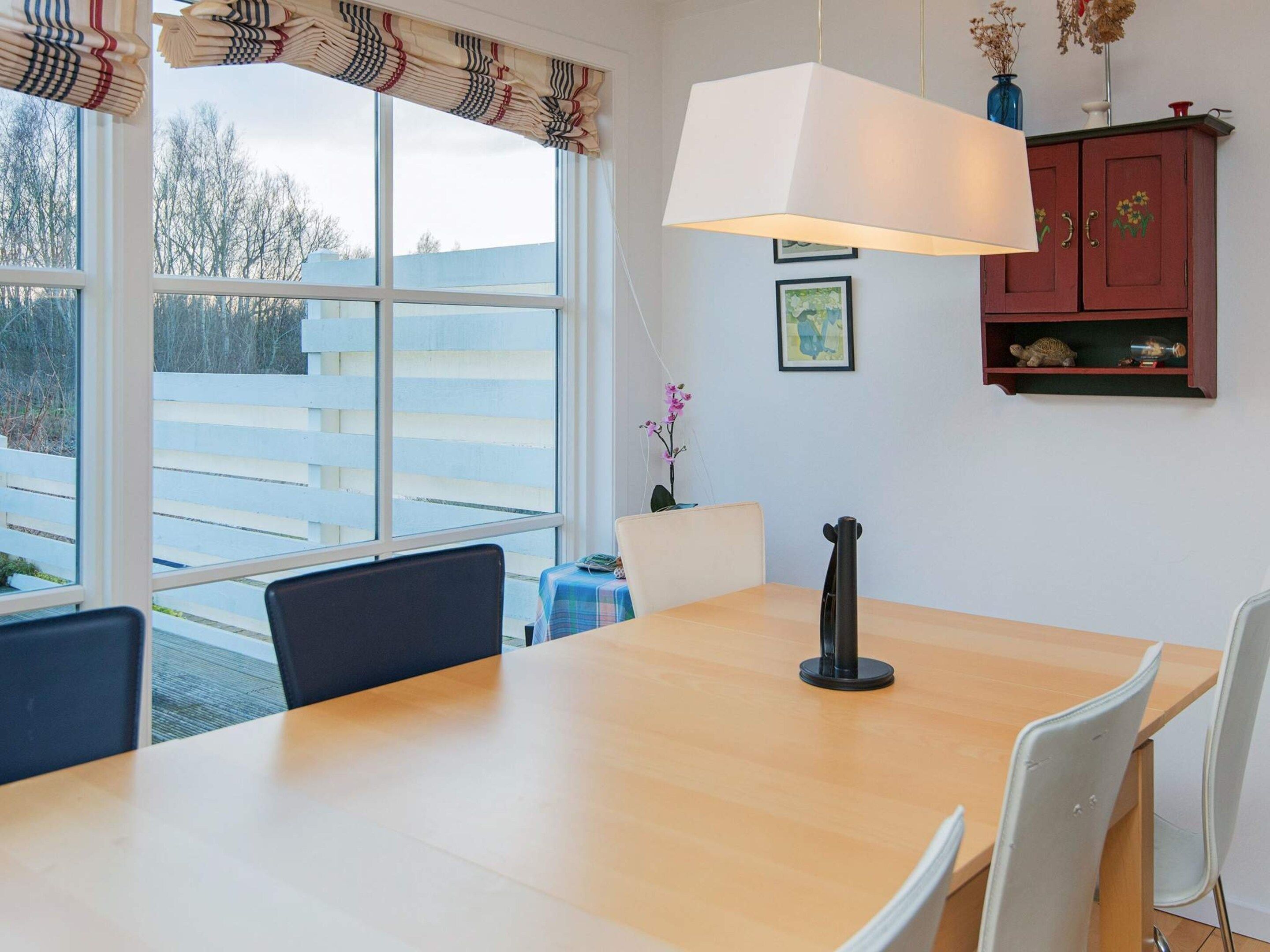 Un comedor con mesa de madera, sillas blancas, una lámpara colgante y vistas al exterior a través de amplios ventanales.