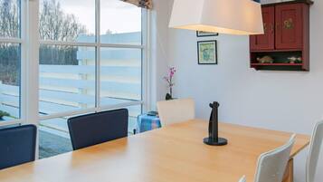 Un comedor con mesa de madera, sillas blancas, una lámpara colgante y vistas al exterior a través de amplios ventanales.