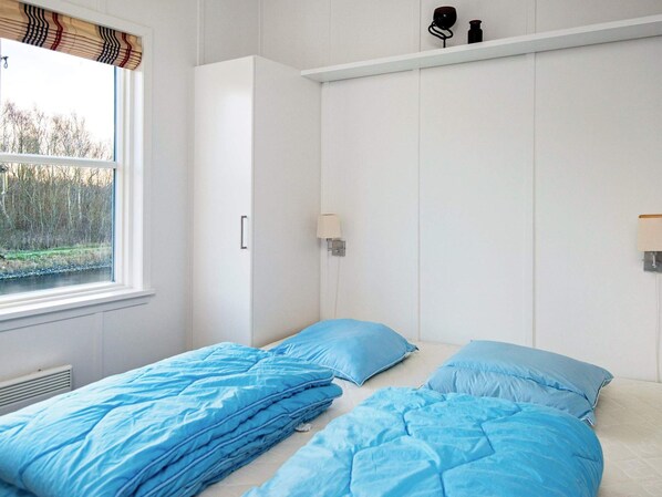 Un dormitorio con dos camas, una ventana que da a árboles, un armario blanco y una estantería con objetos.