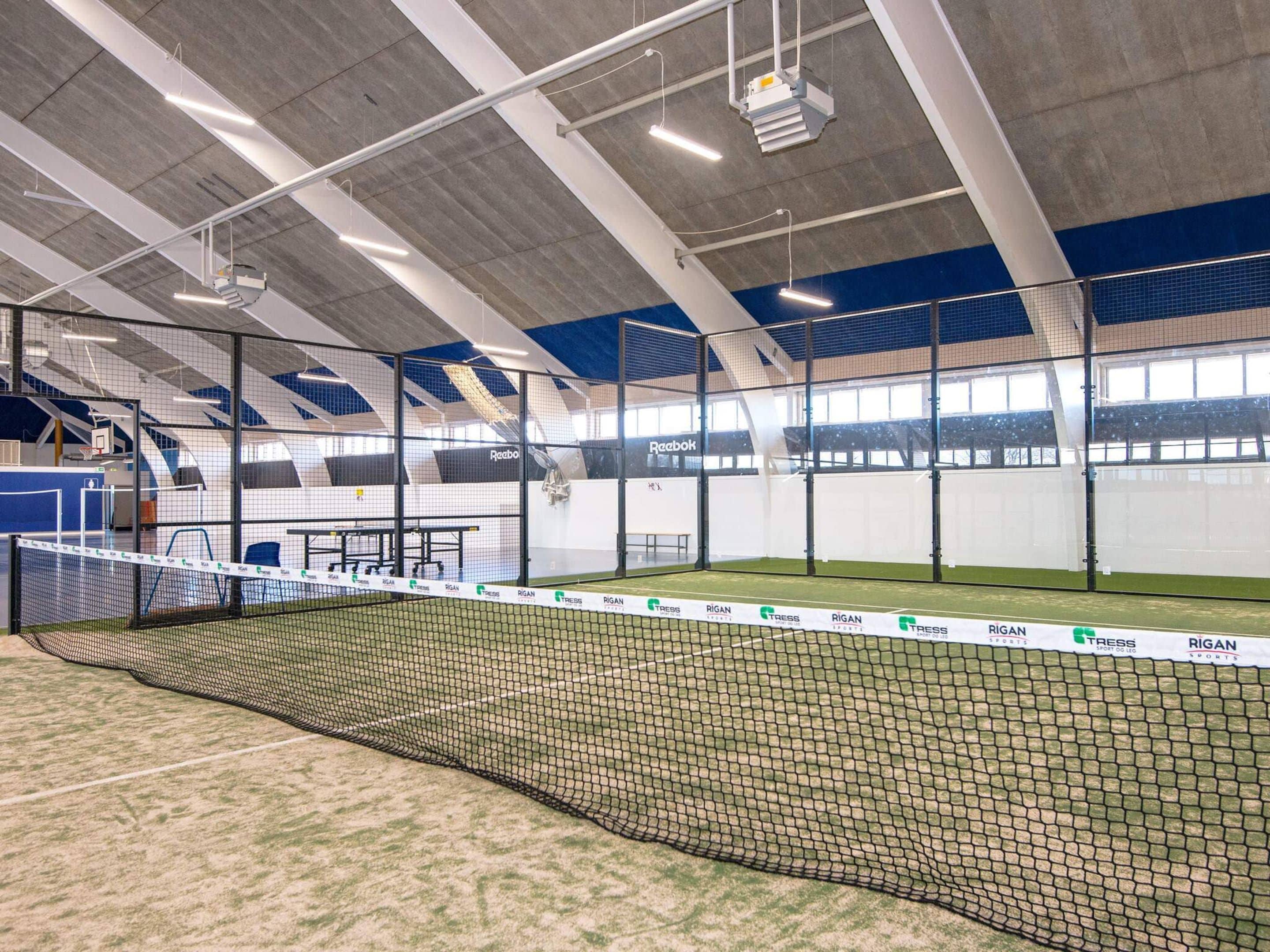 Instalación deportiva interior con cancha de tenis, zonas de estar y un techo alto con luminarias.
