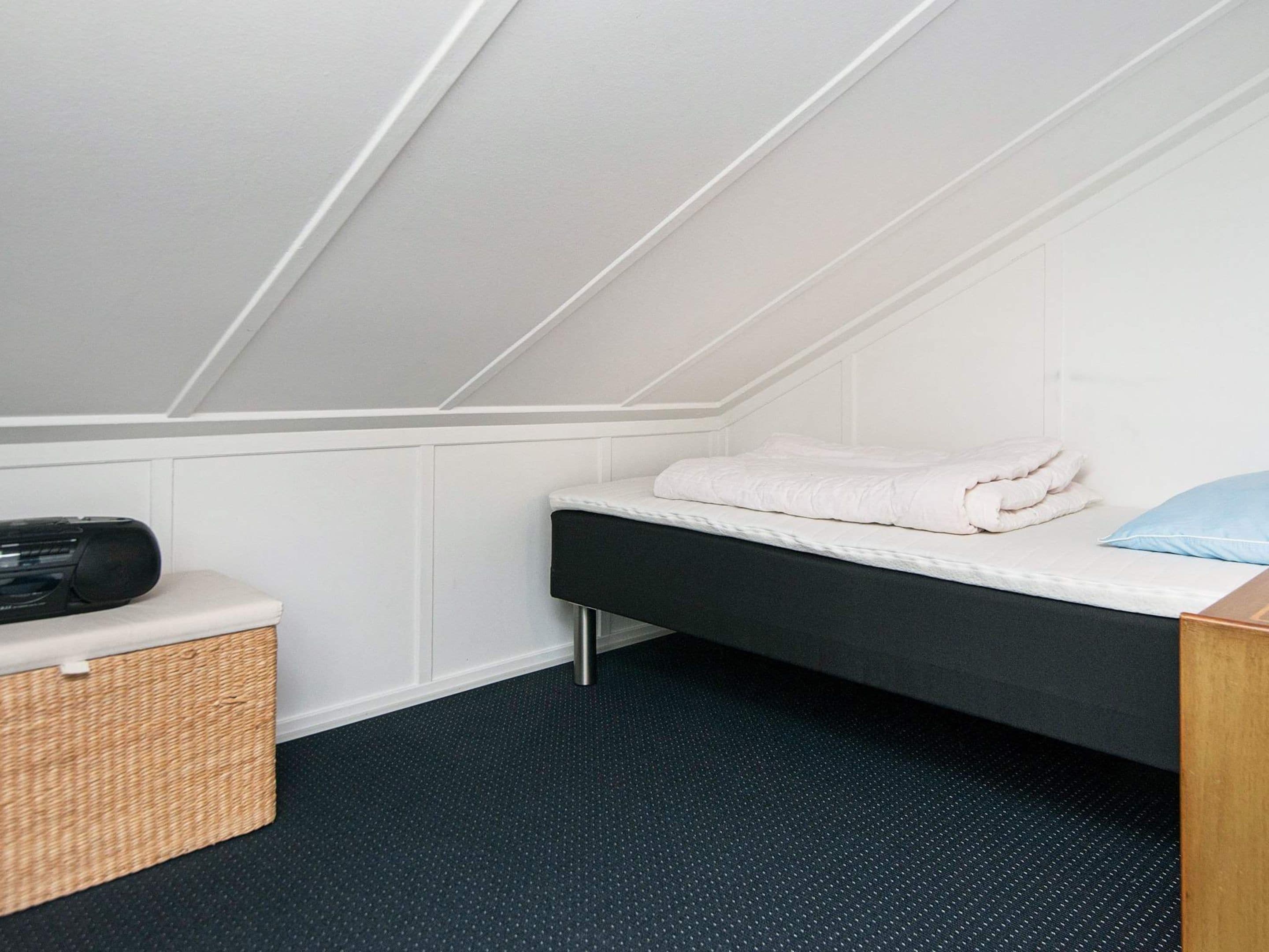 A small, single-bed room with a sloped ceiling, a bedside table with a radio, a basket, and a blue pillow.