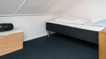 A small, single-bed room with a sloped ceiling, a bedside table with a radio, a basket, and a blue pillow.
