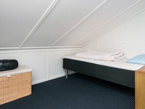 Una habitación pequeña con cama individual, techo inclinado, mesita de noche con radio, una cesta y almohada azul.