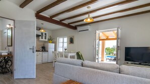 Ein modernes Wohnzimmer mit einer Couch, einem Holztisch und einem Fernseher. Es gibt eine Küchenzeile mit Schränken und einem Kühlschrank. Das Zimmer verfügt über Holzbalken an der Decke und ein großes Fenster.