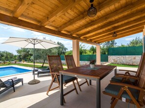 Een terras buiten met een houten tafel en stoelen, een zwembad en uitzicht op het omliggende groen.