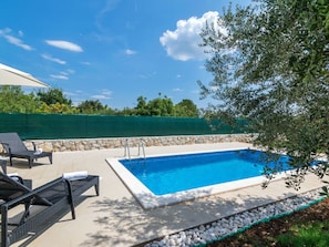 Une piscine rectangulaire bordée de pierres, entourée d’un muret en pierre et d’une clôture verte. On y trouve deux chaises longues avec des coussins, et une serviette est posée sur l’une d’elles.