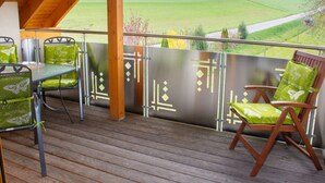 A covered outdoor seating area with a wooden deck, metal chairs, and a view of a green landscape.
