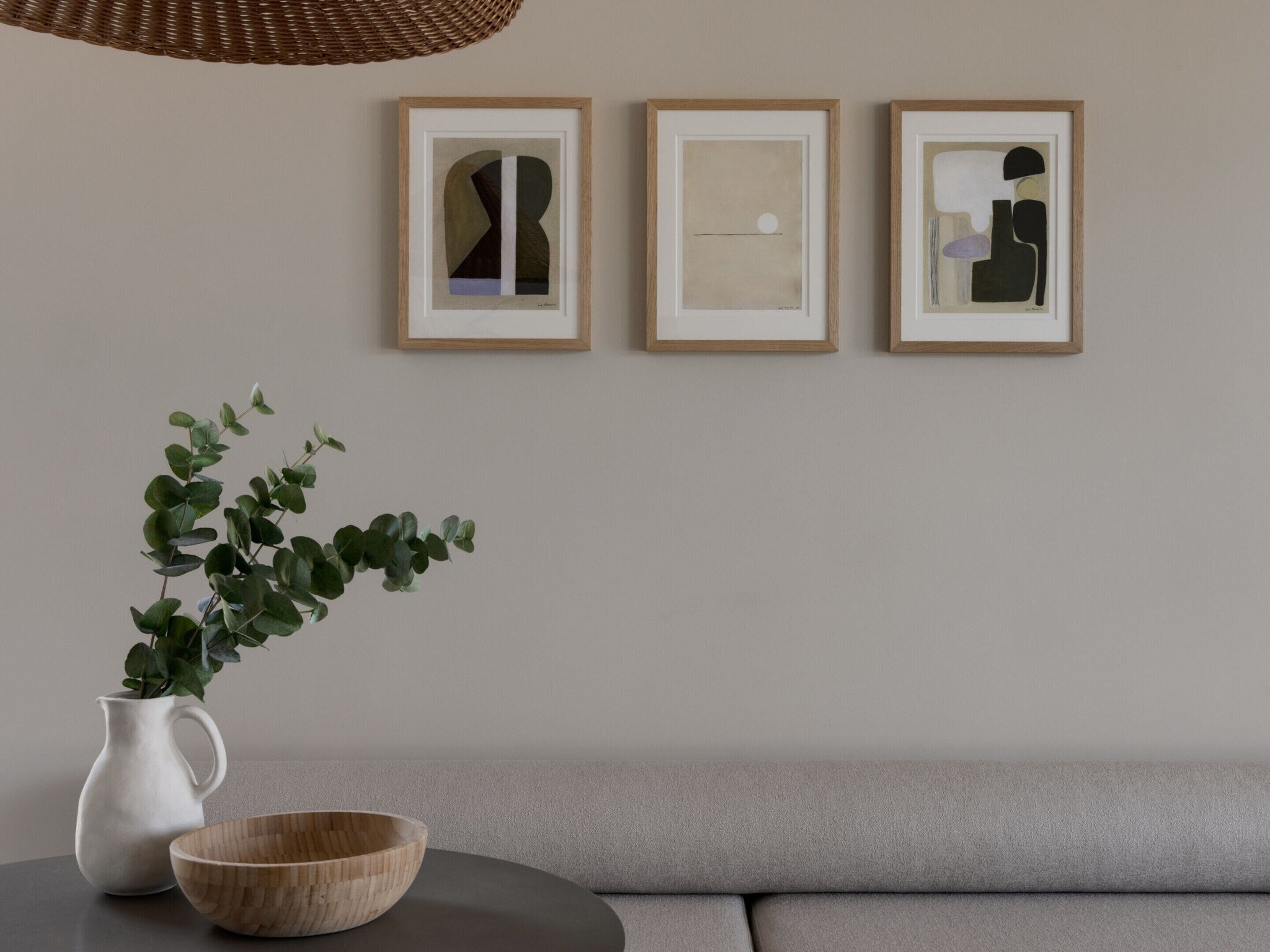 A modern living room with a grey sofa, a round wooden table with a white pitcher and a wooden bowl, and three framed abstract artworks on the wall.
