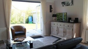 A living room with a glass coffee table, a black leather sofa, a white cabinet, a television, and a wicker chair. - Nice Holiday Home With Garden Near Beach (Wemeldinge)