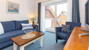 Ett hotellrum med en blå soffa, ett träbord, ett litet sidobord med en lampa och utsikt över ett hus genom en glasdörr.