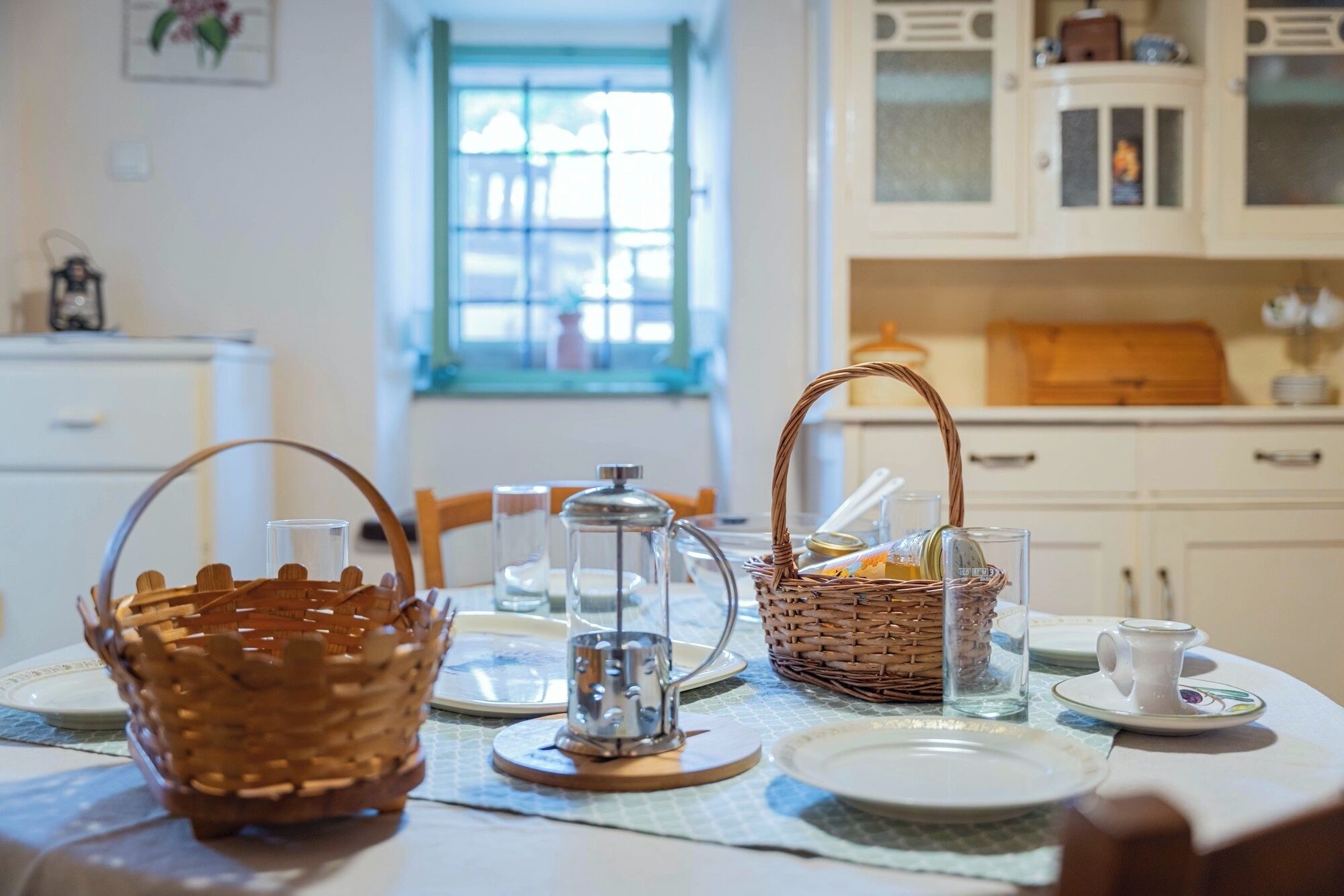 A dining table set with baskets, glasses, and a teapot in a bright kitchen with white cabinets and a window.