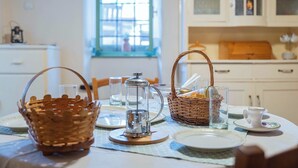 Una mesa de comedor dispuesta con cestas, vasos y una tetera en una cocina luminosa con armarios blancos y un ventanal.
