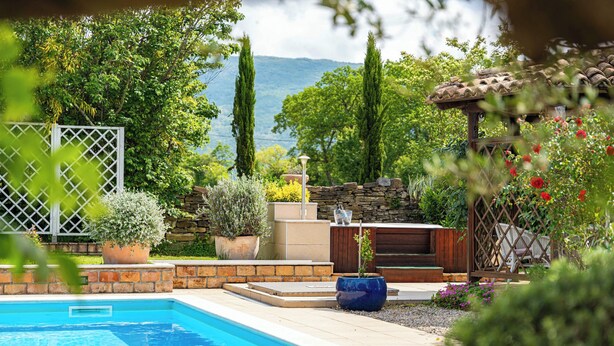 Una piscina rodeada de muros de piedra, plantas en macetas y una pérgola con flores.