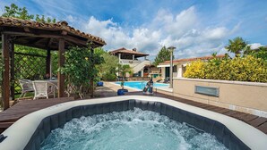 Una bañera de hidromasaje con un muelle de madera y sillas, una piscina, y un edificio con techo de tejas al fondo.