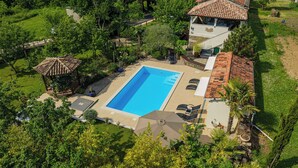 Vista aérea de un resort con piscina rectangular rodeada de sillones y sombrillas, un pabellón y abundante follaje.