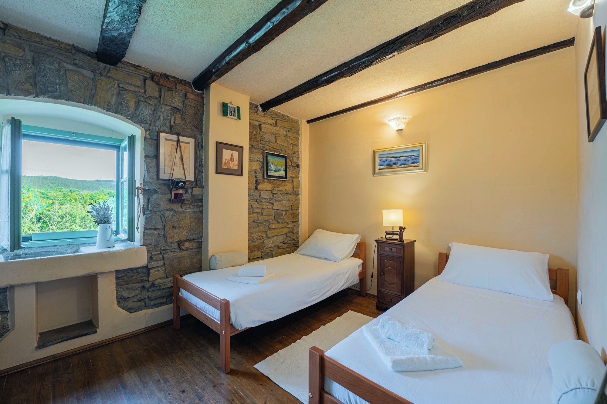 A bedroom with two beds, a stone wall, wooden beams, and a window with a view of greenery.