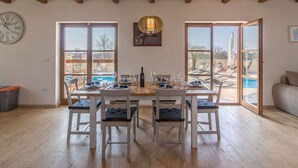 A dining area with a wooden table set for four, surrounded by white chairs with blue cushions. Large windows provide a view of an outdoor swimming pool and lounge chairs. - Villa Malena Comfortable Holiday Residence (Svetvincenat)
