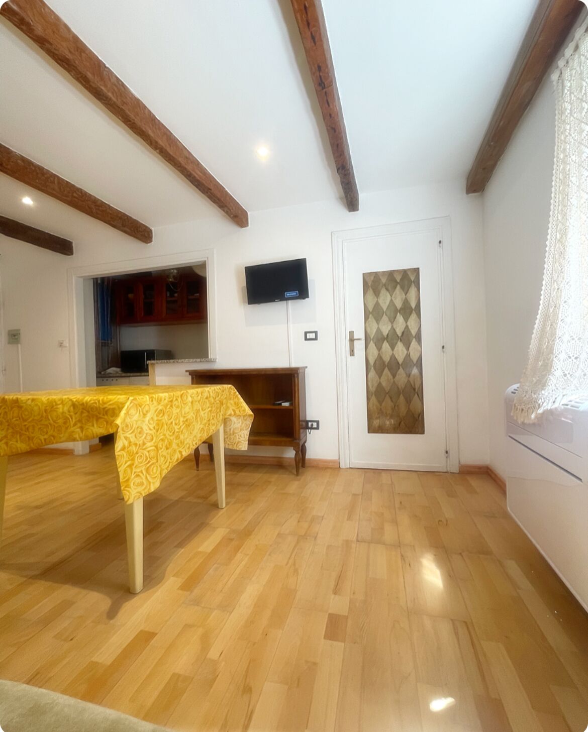 Une pièce avec un plancher en bois, une table à manger recouverte d’une nappe jaune, un téléviseur fixé au mur et une porte dotée d’un panneau de verre à motifs géométriques.