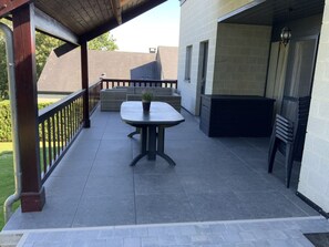 A covered patio with a table and chairs, a small garden area, and a view of a building in the background. - Seaside Comfort for Four (Houffalize)