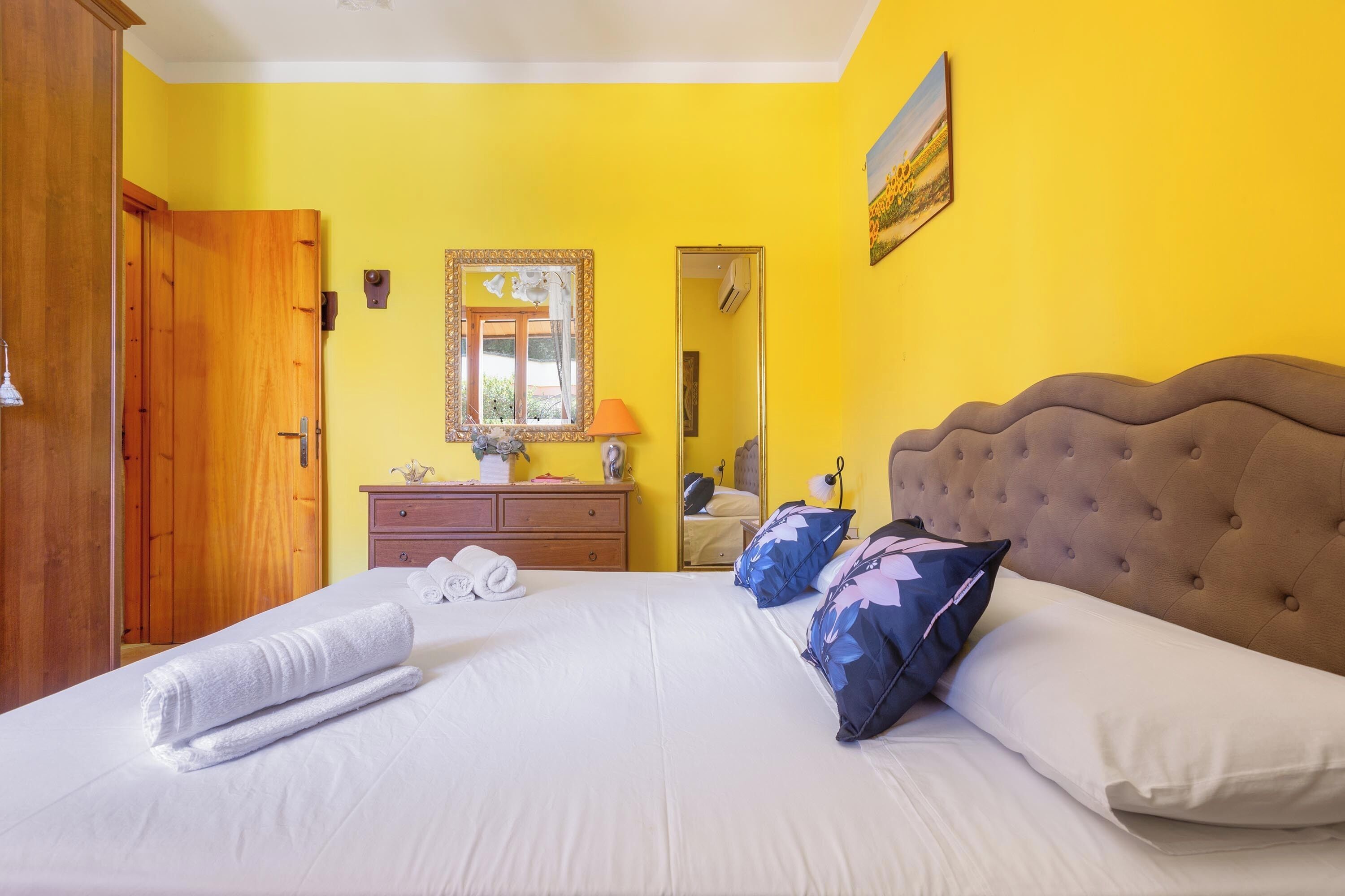 Una cama bien tendida con sábanas blancas y un cabecero marrón, un armario de madera, un aparador con espejo y un cuadro en la pared.