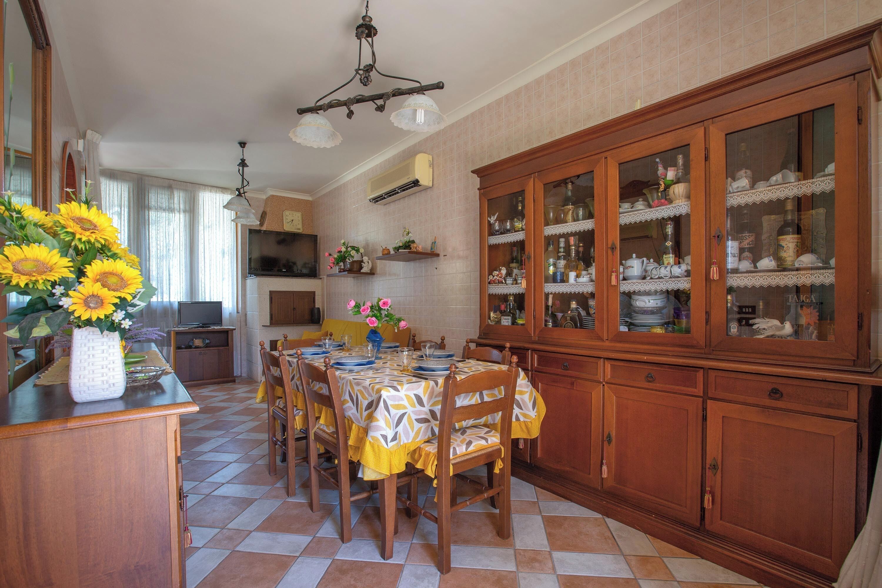 A dining area with a wooden table set for four, surrounded by chairs. There is a cabinet with glass doors to the right, and a vase with sunflowers on the left.