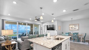 A modern kitchen with a central island, a dining area with teal chairs, and a view of the outdoors. - Sunset FantaSea at One Particular Harbour 380-202 · Sound Views · 3BR (Bradenton)