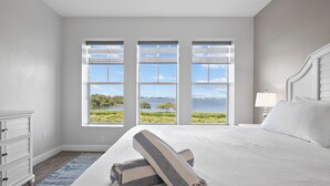 A bedroom with a large bed, a dresser, and a view of the sea through three windows. - Sunset FantaSea at One Particular Harbour 380-202 · Sound Views · 3BR (Bradenton)