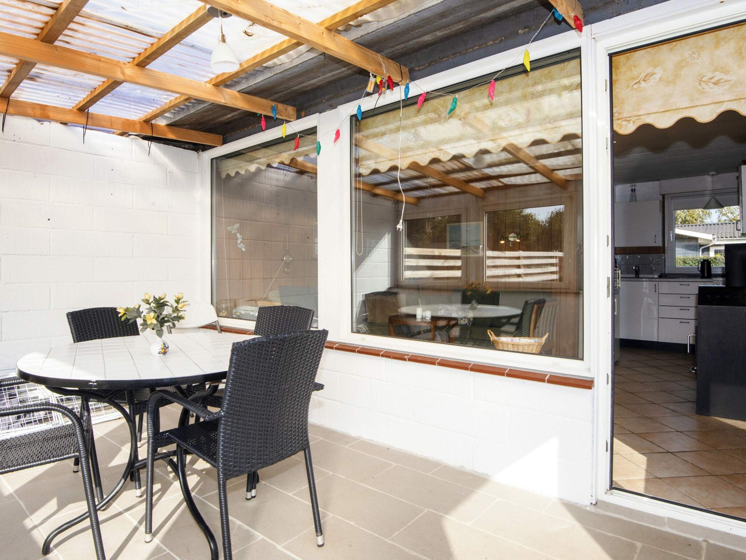 An outdoor patio with a table and chairs, a view of the interior through large windows, and a wooden ceiling structure.