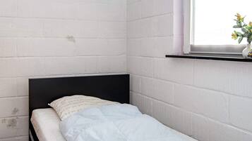 A small, single-bed room with white brick walls, a black headboard, and a window with a plant on the sill.