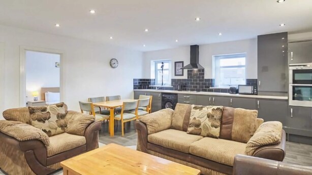 A modern living room with two sofas, a wooden coffee table, and a dining table with chairs. A kitchen with a stove and cabinets is visible in the background. - Jackson Court Apartment 3 (Redcar)